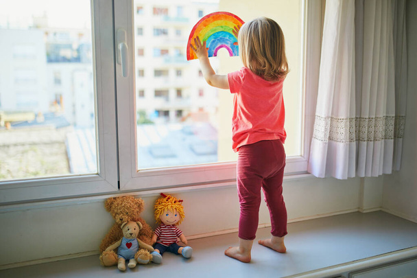 Adorable toddler girl attaching rainbow drawing to window glass as sign of hope. Creative games for kids staying at home during lockdown. Self isolation and coronavirus quarantine concept - Фото, изображение