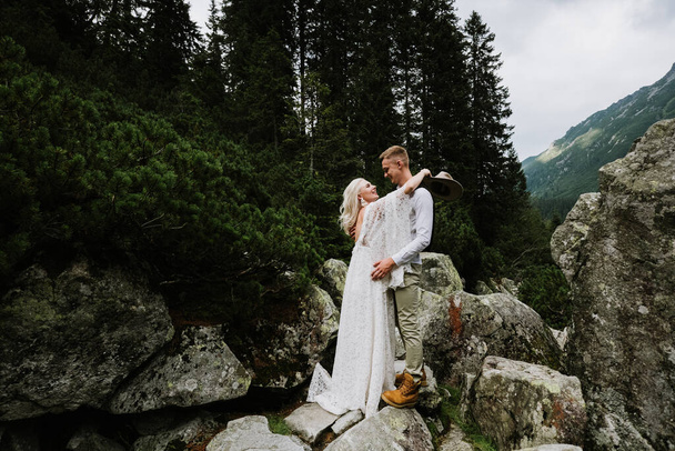Όμορφη νύφη σε boho στυλ φόρεμα, καπέλο και γαμπρός αγκαλιάζονται στα βουνά. - Φωτογραφία, εικόνα