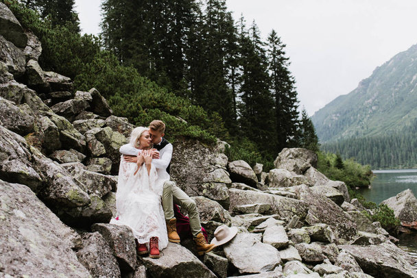 Όμορφη νύφη σε boho στυλ φόρεμα και αγκαλιά γαμπρός κάθεται σε πέτρες δίπλα στη λίμνη στα βουνά. - Φωτογραφία, εικόνα
