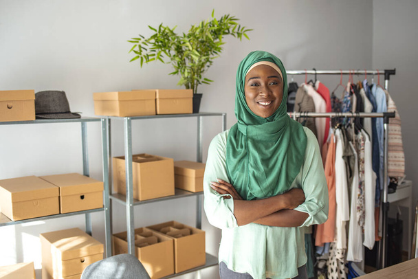 Muslim woman designer in her tailor shop. Happy smiling muslim woman designer as a startup business owner working in her tailor shop. Young women in office, looking at camera and smiling - 写真・画像