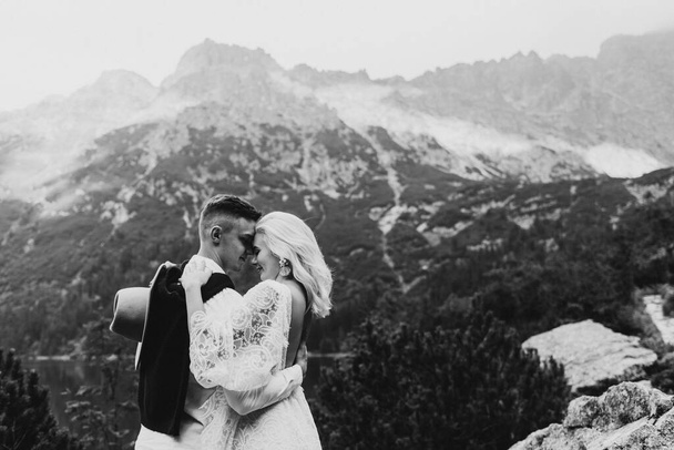Boho tarzı elbiseli güzel bir gelin, dağlarda şapka ve damat sarılıyor. Dağlarda bir gelin ve damadın siyah beyaz fotoğrafı. - Fotoğraf, Görsel
