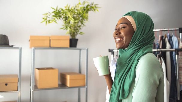 A Middle-eastern woman is indoors in her home office. She is wearing a head scarf. She is smiling behind her while sitting at her desk. She has coffee mugs in her hand.  - Photo, image