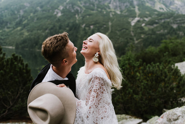 Mooie bruid in een boho stijl jurk, hoed en bruidegom knuffel en lach in de bergen. - Foto, afbeelding