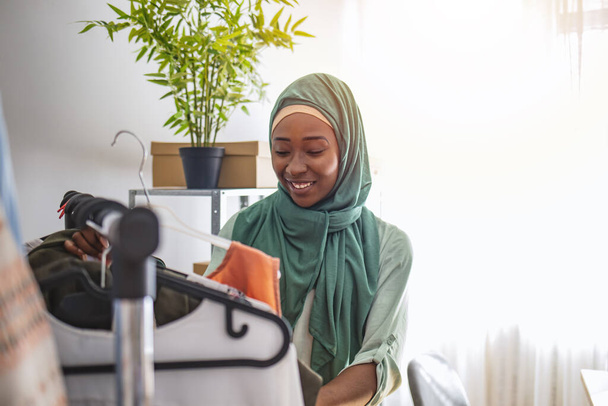 Everyone do shopping online. Woman packing item that she sells online. Muslim woman designer as a startup business owner. Female muslim designer checking hanged clothes in tailor shop  - 写真・画像