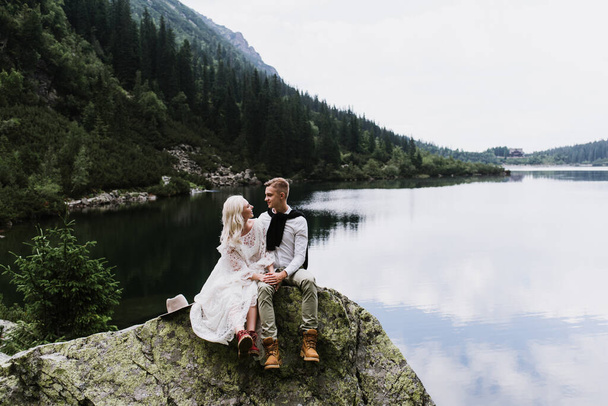 Boho tarzı bir elbise içinde güzel bir gelin. Şapka ve damat dağlardaki bir gölün kenarında taşların üzerinde oturuyor.. - Fotoğraf, Görsel