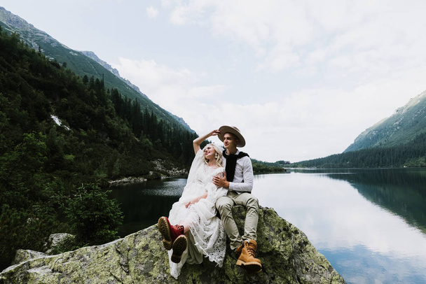 Beautiful bride in a boho style dress, hat and groom are sitting on stones near a lake in the mountains. - Photo, Image