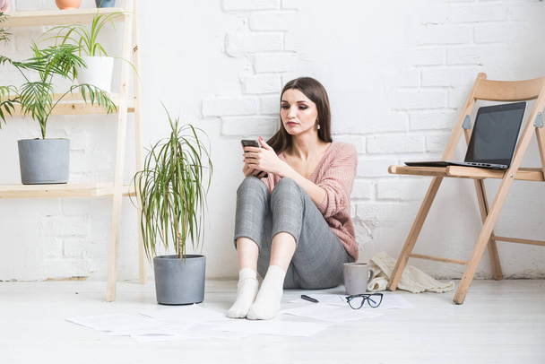 A happy woman in homemade casual clothes sits in a bright interior and uses a telephone, the girl is resting while working at a laptop and surfing the Internet. Cozy freelance work space, - 写真・画像