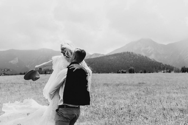 Der Bräutigam umkreist die Braut in den Bergen auf dem Arm. Der Bräutigam hält die Braut in einem Boho-Kleid und einem Hut in den Händen in den Bergen. Die Braut schwenkt ihren Hut in den Bergen. - Foto, Bild
