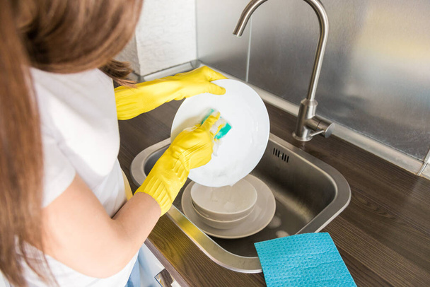 A young woman in yellow gloves washes dishes with a sponge in the sink. House professional cleaning service. - Φωτογραφία, εικόνα