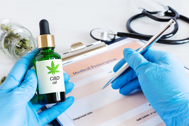 Doctor prescribing CBD oil for patient. Bottle of Cbd oil, cannabis seeds and dry leaves over medical prescription sheet top view on white wooden table. Medical marijuana, alternative medicine concept. - Foto, afbeelding