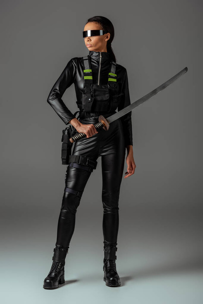 attractive futuristic african american woman in glasses with sword on grey - Фото, изображение