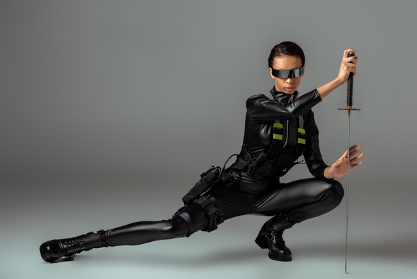 aantrekkelijk futuristisch Afrikaans amerikaans vrouw in bril zitten met zwaard op grijs - Foto, afbeelding