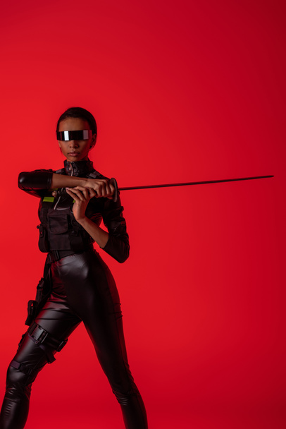 vonzó futurisztikus afro-amerikai nő szemüvegben karddal elszigetelt vörös háttér - Fotó, kép