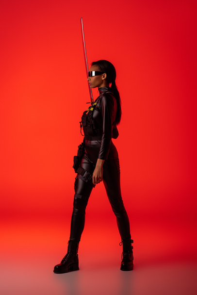 attractive futuristic african american woman in glasses with sword on red background - Valokuva, kuva