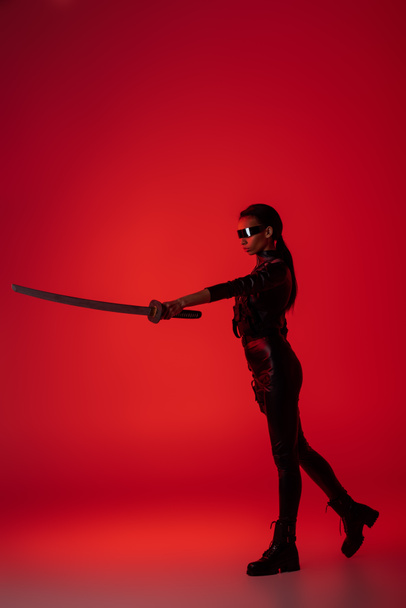 attraktive, futuristische afrikanisch-amerikanische Frau in Brille mit Schwert auf rotem Hintergrund - Foto, Bild