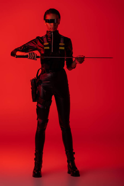 attractive futuristic african american woman in glasses with sword on red background - 写真・画像