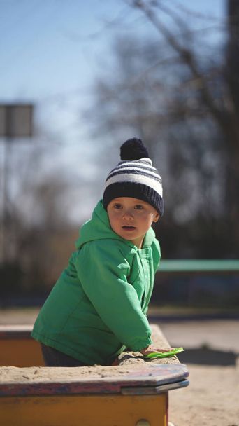 公園の遊び場のサンドボックスの緑のジャケットの少年。子供は目を背けて - 写真・画像