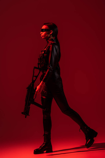 futurisztikus afro-amerikai nő szemüvegben séta támadás puska piros háttér - Fotó, kép
