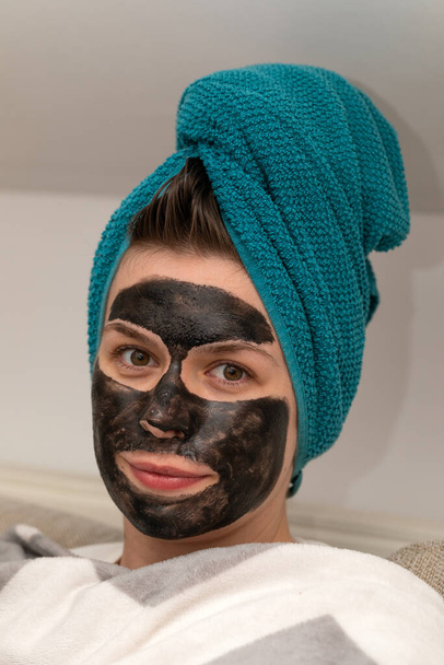 Portrait of woman having a black charcoal face mask on, with towel on head, waiting for the results at home - Photo, Image