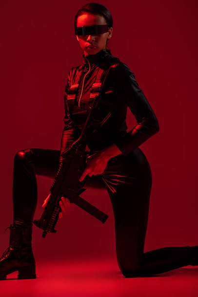 futuristic african american woman in glasses standing on knee with assault rifle on red background - Photo, Image