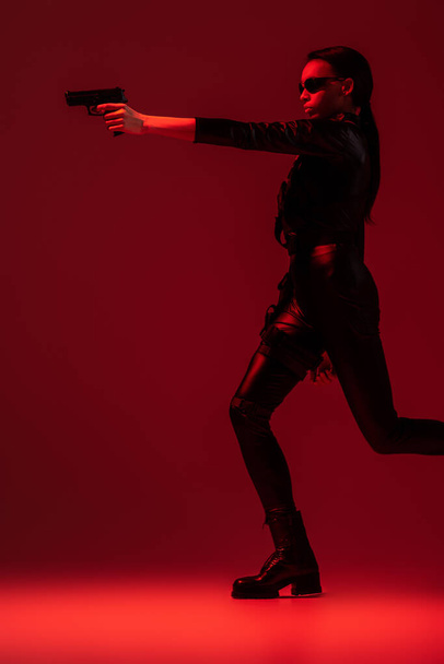 futuristic african american woman in glasses aiming gun on red background - 写真・画像