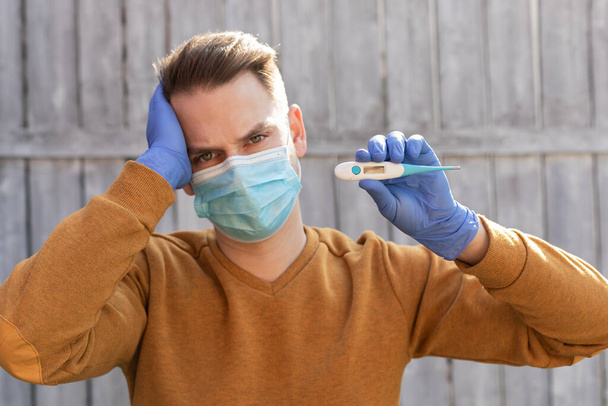 Portrait of a young man contaminated with covid-19 is wearing mask and gloves, holding a digital thermometer  - Foto, Bild