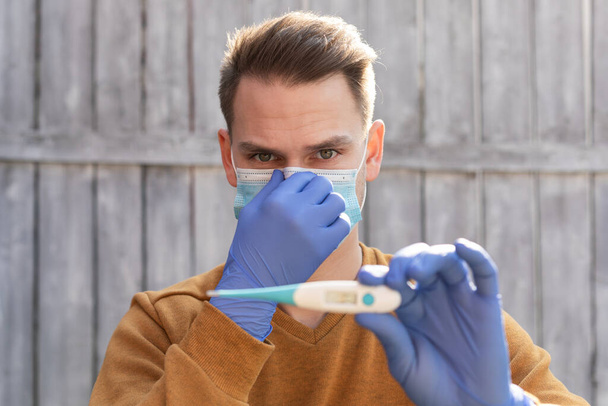 Portrait of a young man contaminated with covid-19 is wearing mask and gloves, holding a digital thermometer  - Photo, Image