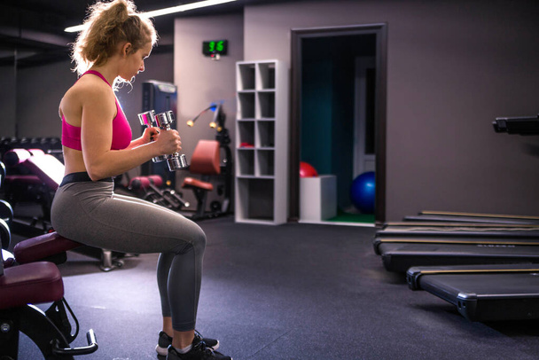 Girl posing in the gym with dumbbells in hands, sitting on a bench. Horizontal photo - Photo, Image