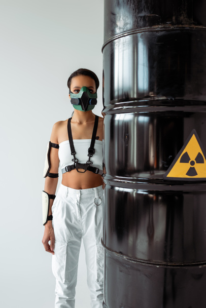 futuristic african american woman in safety mask near radioactive waste barrels isolated on white - Foto, Imagem