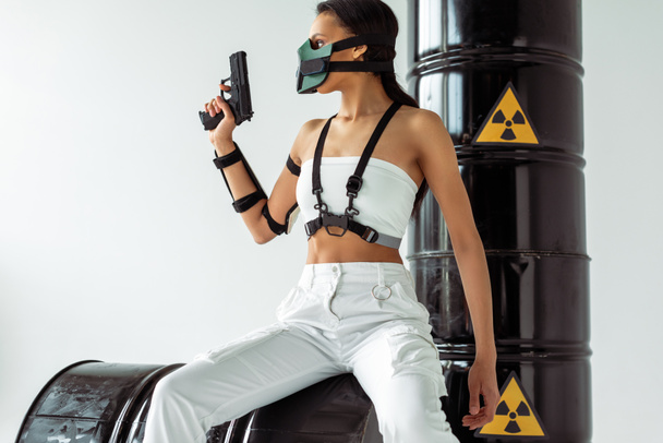 futurista mujer afroamericana en máscara de seguridad con arma cerca de barriles de residuos radiactivos aislados en blanco
 - Foto, imagen