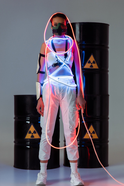 mulher africana futurista americana em máscara de segurança e luz de néon perto de barris de resíduos radioativos
 - Foto, Imagem