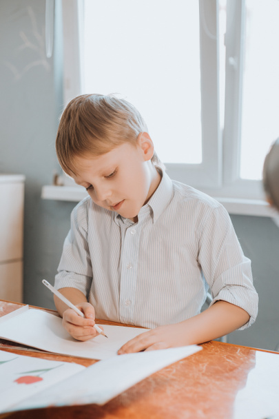 Chłopiec pisze w notatniku w domu podczas kwarantanny CoVid-19, nauka na odległość online z laptopem, dziecko odrabiające lekcje do szkoły. Dzieci zostały w domu.. - Zdjęcie, obraz