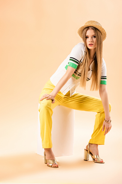 fashionable young woman in yellow trousers, polo and straw hat sitting on white cube on beige - Фото, зображення