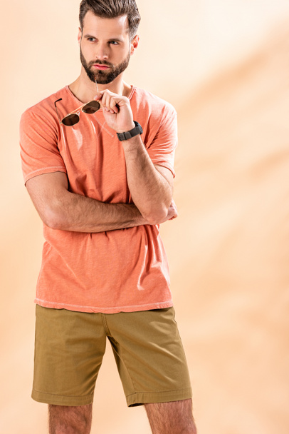 handsome man posing in shorts and summer t-shirt holding sunglasses on beige - Fotoğraf, Görsel