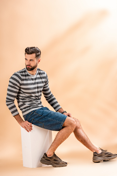 handsome bearded man in striped sweatshirt sitting on white cube on beige - Photo, Image