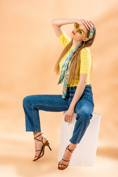menina loira na moda em jeans, camiseta amarela, óculos de sol, sandálias de salto alto e cachecol de seda sentado no cubo branco no bege
 - Foto, Imagem