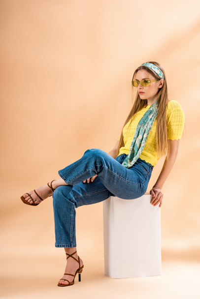módní blondýnka v džínách, žlutém tričku, slunečních brýlích, podpatcích sandálech a hedvábném šátku sedícím na bílé kostce na béžové - Fotografie, Obrázek