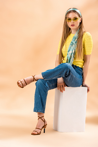 stylová blondýnka v džínách, žlutém tričku, slunečních brýlích, podpatcích sandálech a hedvábném šátku sedícím na bílé kostce na béžové - Fotografie, Obrázek
