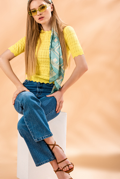linda menina loira em jeans, camiseta amarela, óculos de sol, sandálias de salto alto e cachecol de seda sentado no cubo branco em bege
 - Foto, Imagem