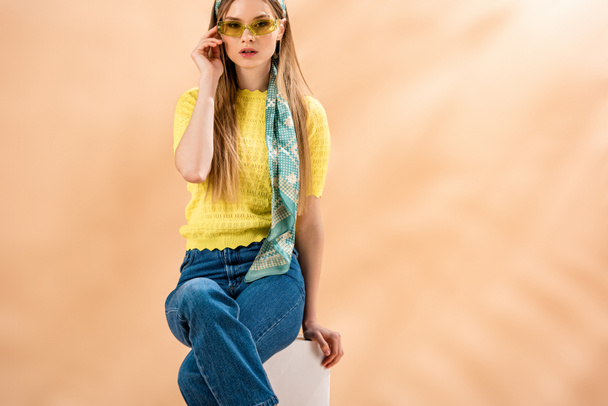 fille à la mode en jeans, t-shirt jaune, lunettes de soleil et foulard en soie assis sur cube blanc sur beige
 - Photo, image