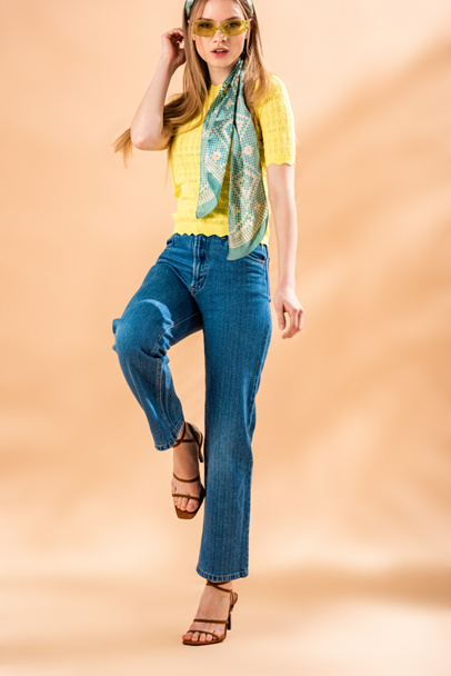 atraktivní dívka pózující v džínách, žlutém tričku, slunečních brýlích a hedvábném šátku na béžové - Fotografie, Obrázek