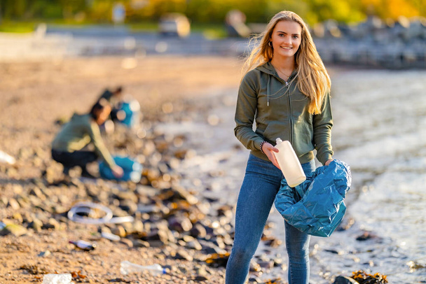 Kumsalda elinde şişe ve çöp torbası tutan genç ve güzel bir gönüllü. - Fotoğraf, Görsel
