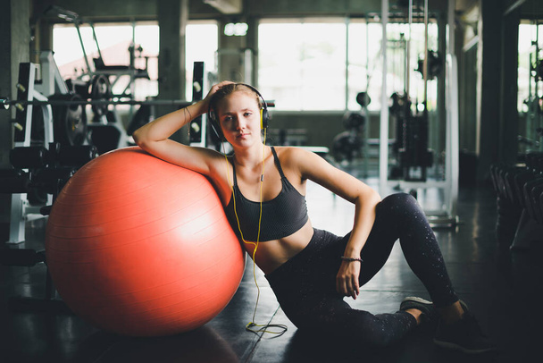 A beautiful woman wearing a sports shirt is sitting and relaxing by listening to music from headphones. With rubber  yoga ball or exercise ball - Photo, image