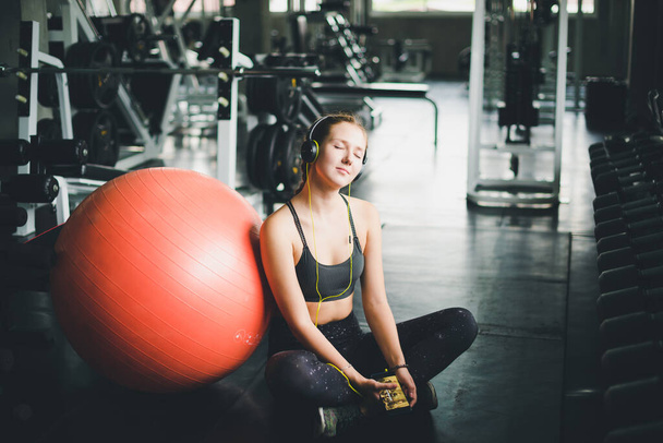 Krásná žena ve sportovní košili sedí a relaxuje posloucháním hudby ze sluchátek. S gumovým jóga míčkem nebo cvičným míčkem - Fotografie, Obrázek