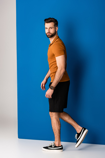 stylish young man posing in shorts and brown polo on grey and blue  - Fotoğraf, Görsel