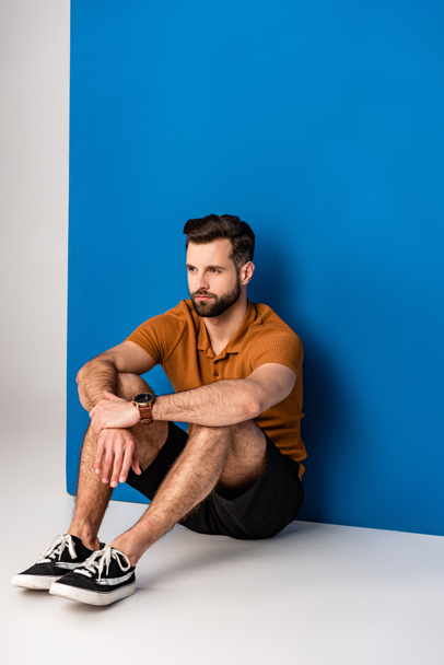 handsome bearded man sitting in shorts and brown polo on grey and blue - Photo, Image