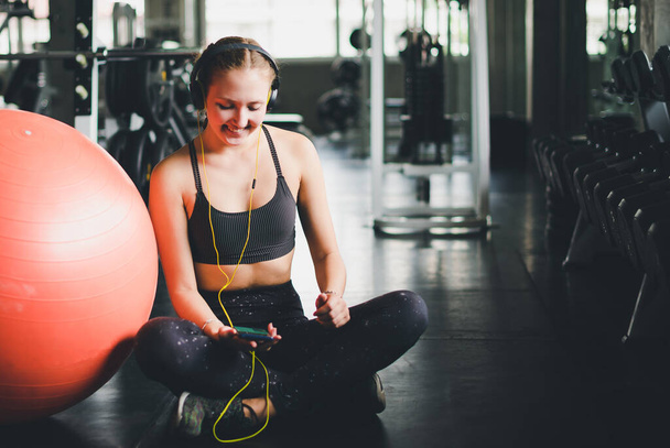A beautiful woman wearing a sports shirt is sitting and relaxing by listening to music from headphones. With rubber  yoga ball or exercise ball - Foto, imagen