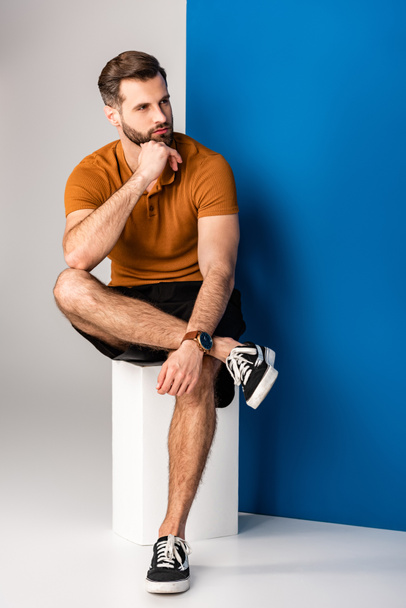 pensive bearded man in shorts and brown polo sitting on white cube on grey and blue - Foto, Imagem