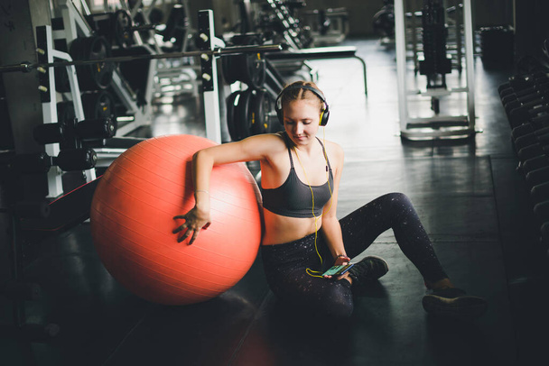 A beautiful woman wearing a sports shirt is sitting and relaxing by listening to music from headphones. With rubber  yoga ball or exercise ball - Fotó, kép
