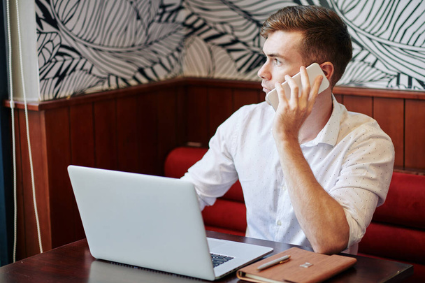 Молодой бизнесмен работает на ноутбуке за столиком кафе, смотрит в окно и разговаривает по телефону с клиентом - Фото, изображение